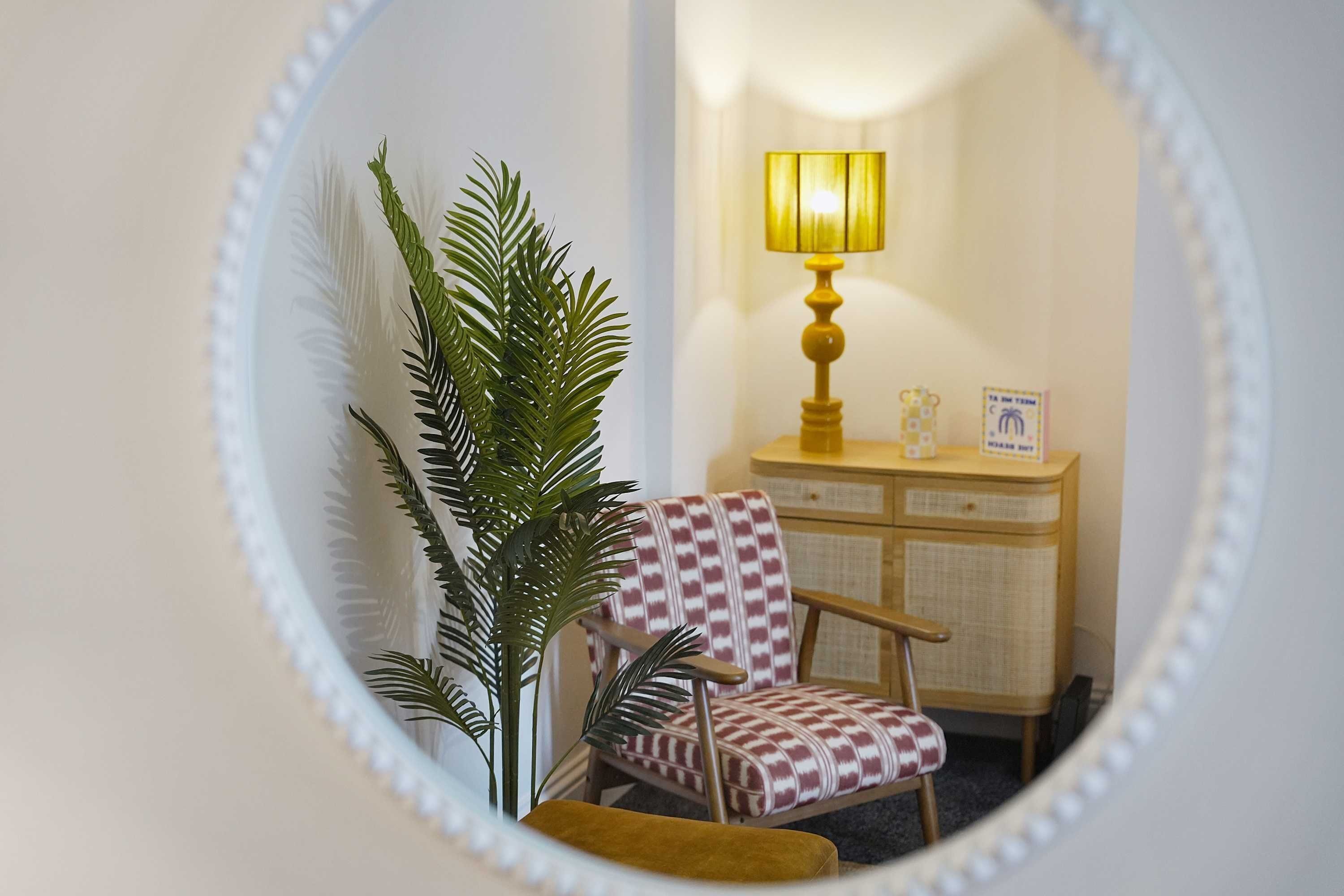 A room with a mirror reflecting a chair, a lamp, and a cabinet.