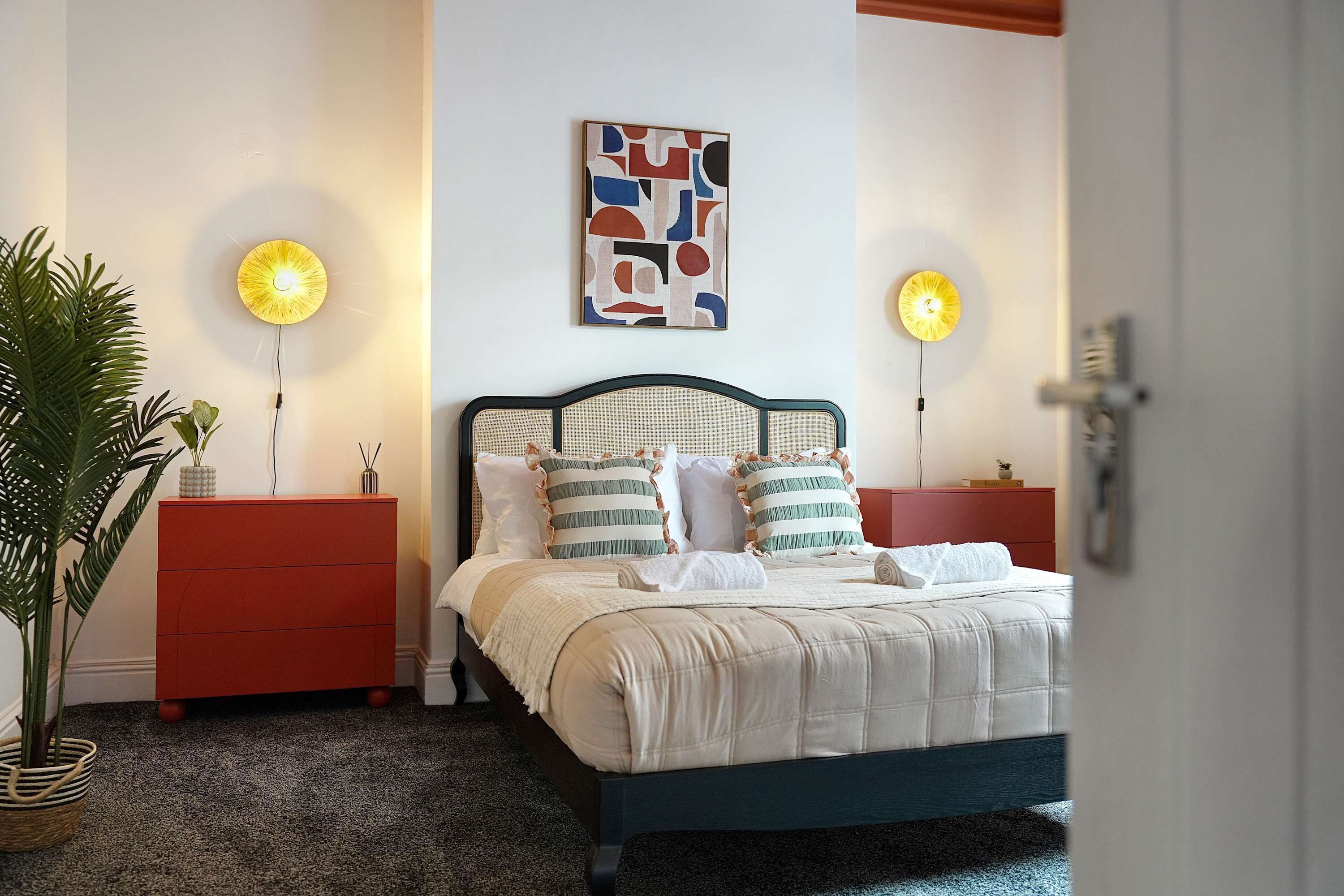 A bedroom with a bed, two bedside tables, a wall art piece, and two wall-mounted lamps.