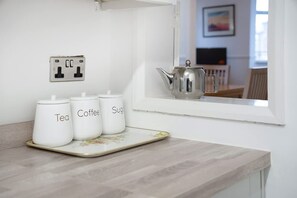Uma bancada de cozinha com três recipientes brancos rotulados 'Tea', 'Coffee' e 'Sugar', e uma chaleira de prata num parapeito.