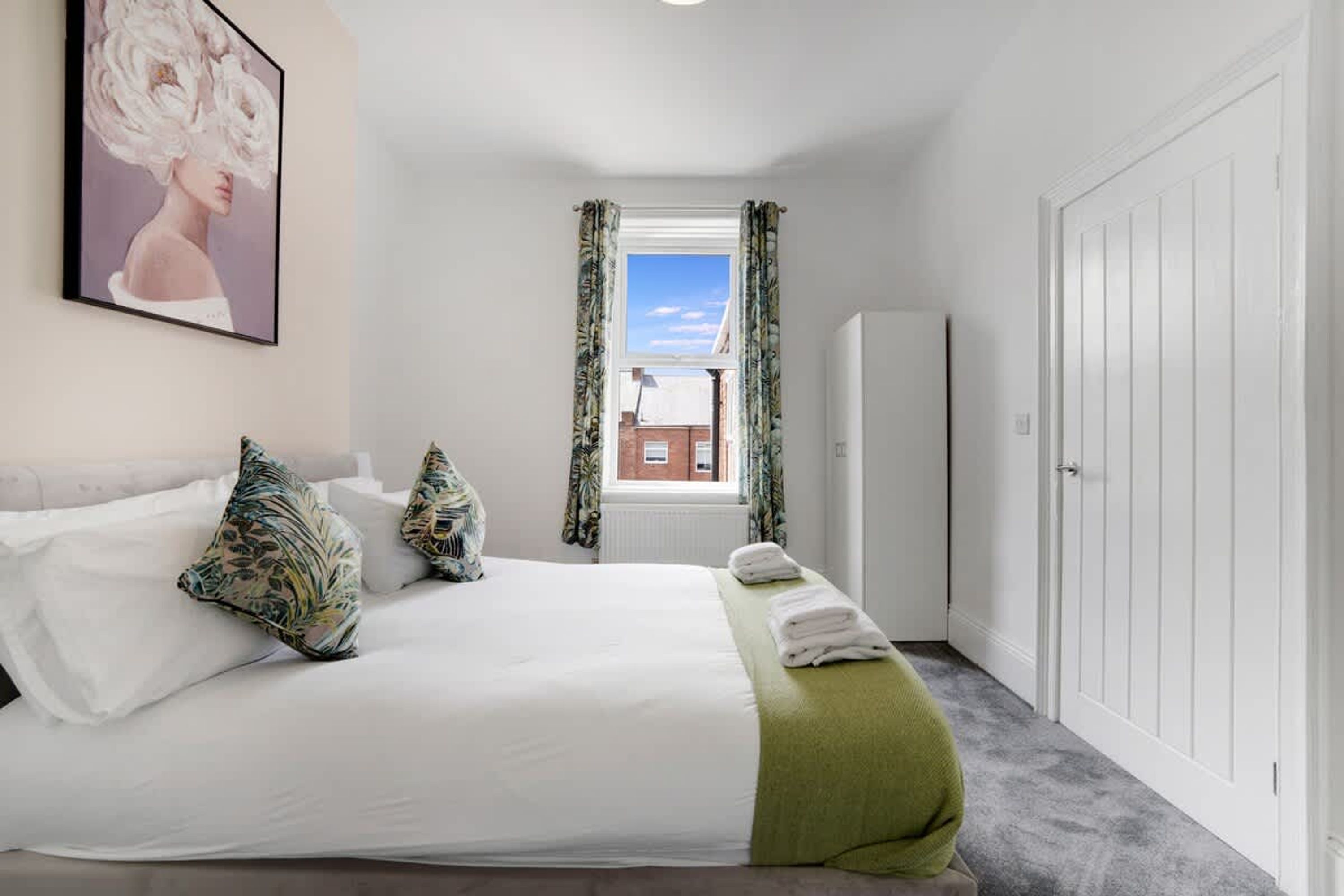 A bedroom with a bed, pillows, a green throw, a window with curtains, a radiator, and a wardrobe.