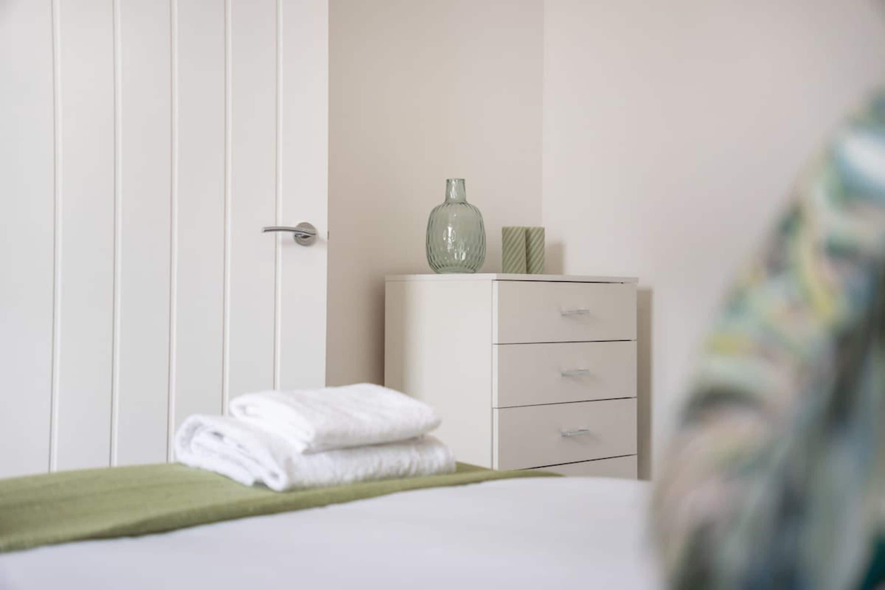 A bedroom with a bed, a green towel, a dresser, and a vase on top.