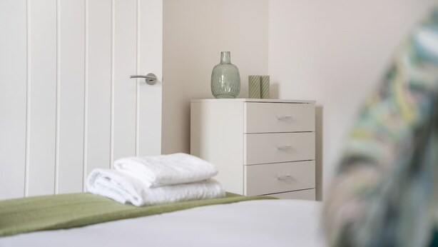 A bedroom with a bed, a green towel, a dresser, and a vase on top. - 4BR South Shields Stay Sleeps 8 by Stay With Us (South Shields)