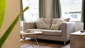 A living room with a sofa, a small round table, and a view of a building through a window. - Stylish 1-bed Stay in the Heart of Barnard Castle (Barnard Castle)