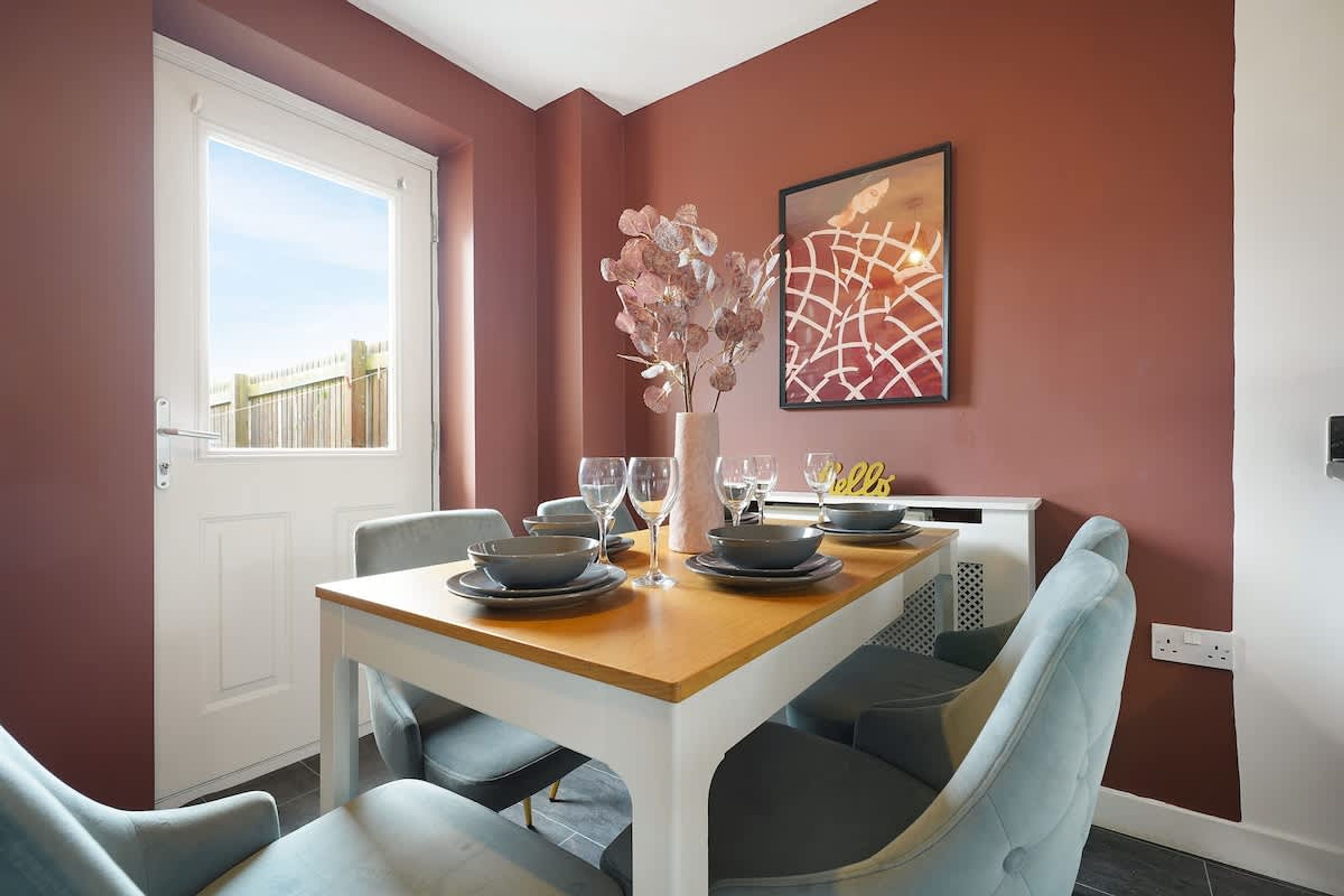 Un coin repas avec une table dressée pour quatre personnes, des chaises, un vase rempli de fleurs et un tableau accroché au mur.