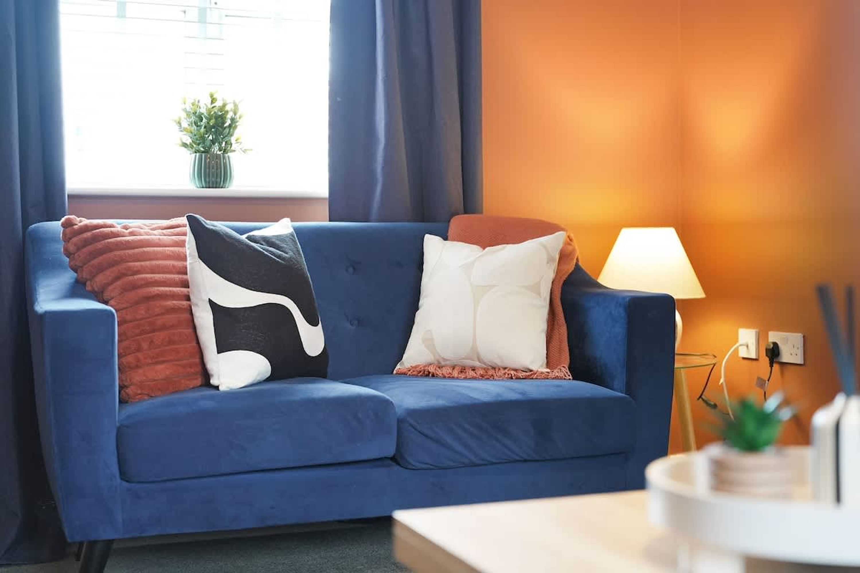 A blue sofa with multiple cushions, a small table with a lamp, and a window with curtains.