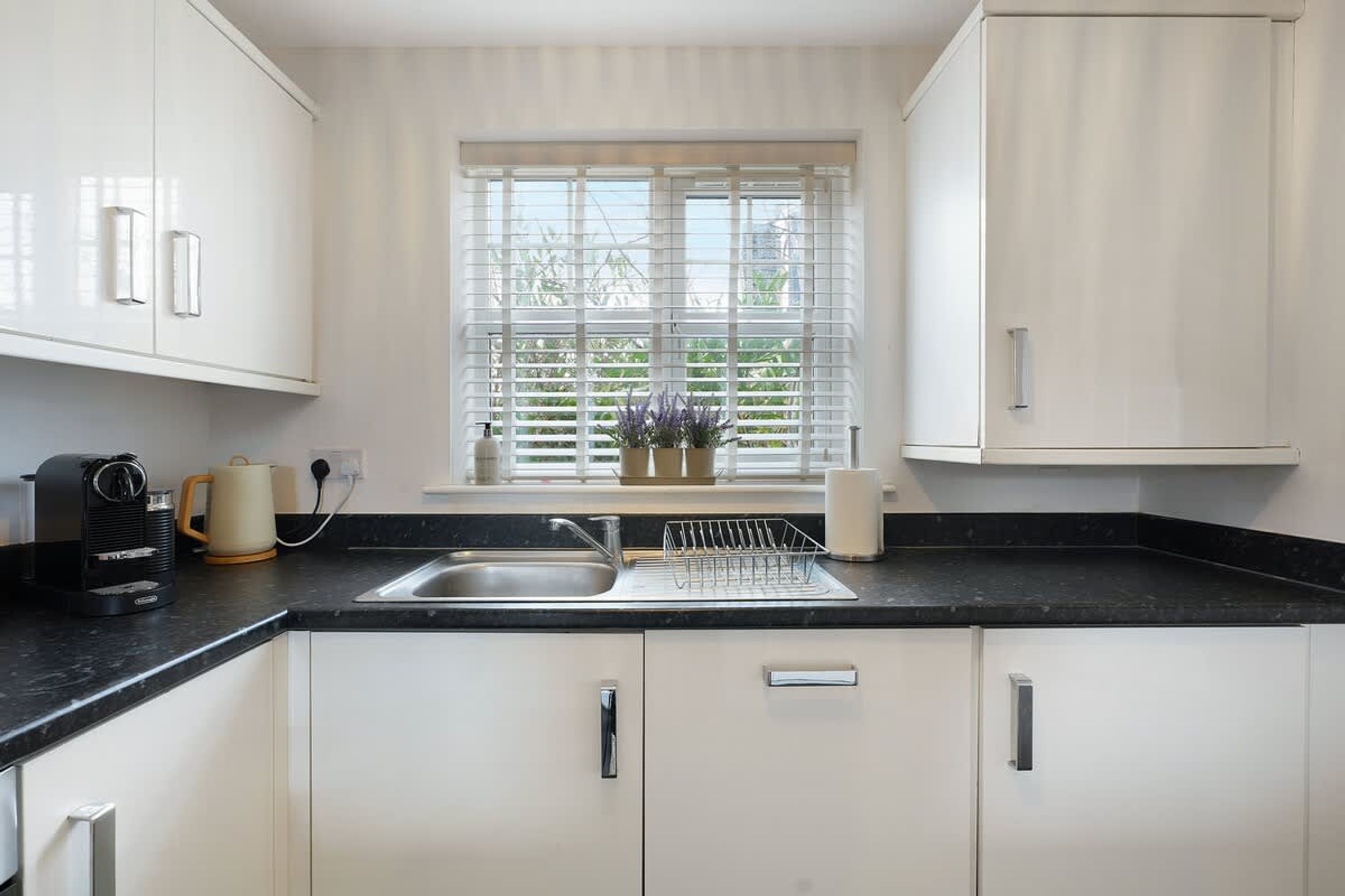 A modern kitchen with white cabinets, a black countertop, a built-in sink, and a window with blinds.
