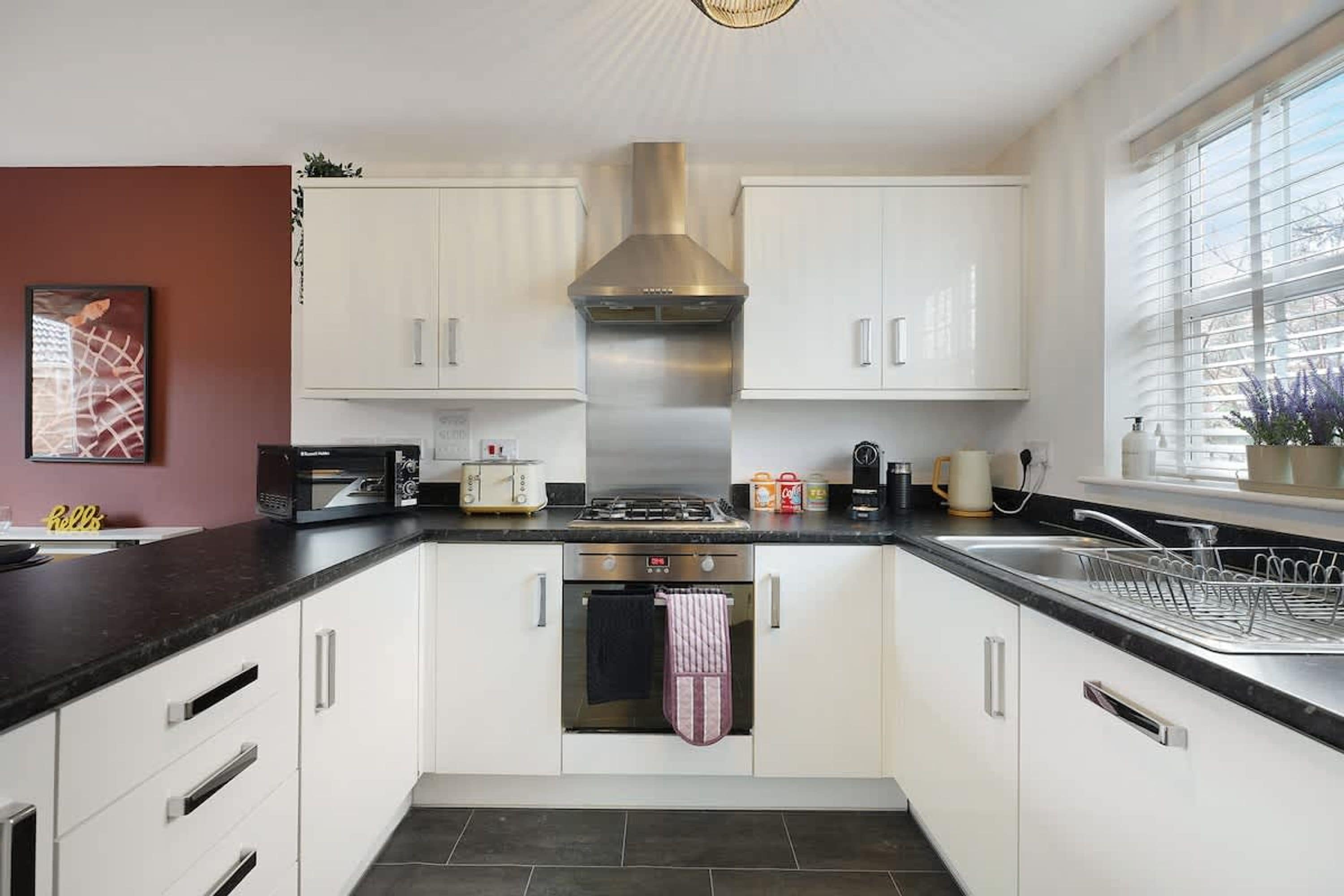 A modern kitchen with white cabinets, a stainless steel range hood, a built-in oven, and a window with blinds.