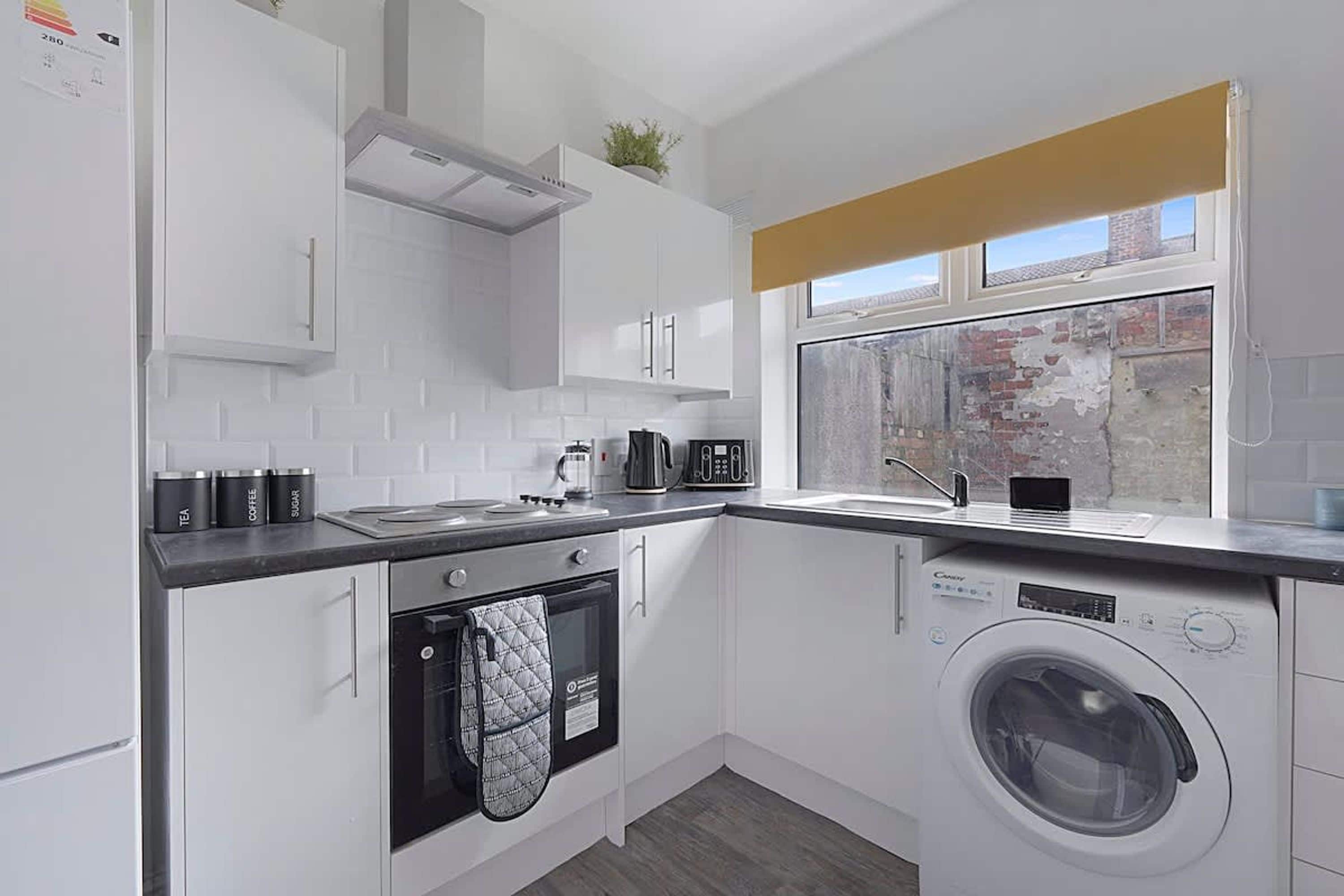 Une cuisine compacte avec des meubles blancs, un four encastré, un lave-linge et une fenêtre donnant sur un mur de briques.