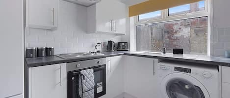 A compact kitchen with white cabinets, a built-in oven, a washing machine, and a window with a view of a brick wall.