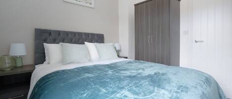 A bedroom with a large bed, a nightstand, a lamp, a wardrobe, and a door.