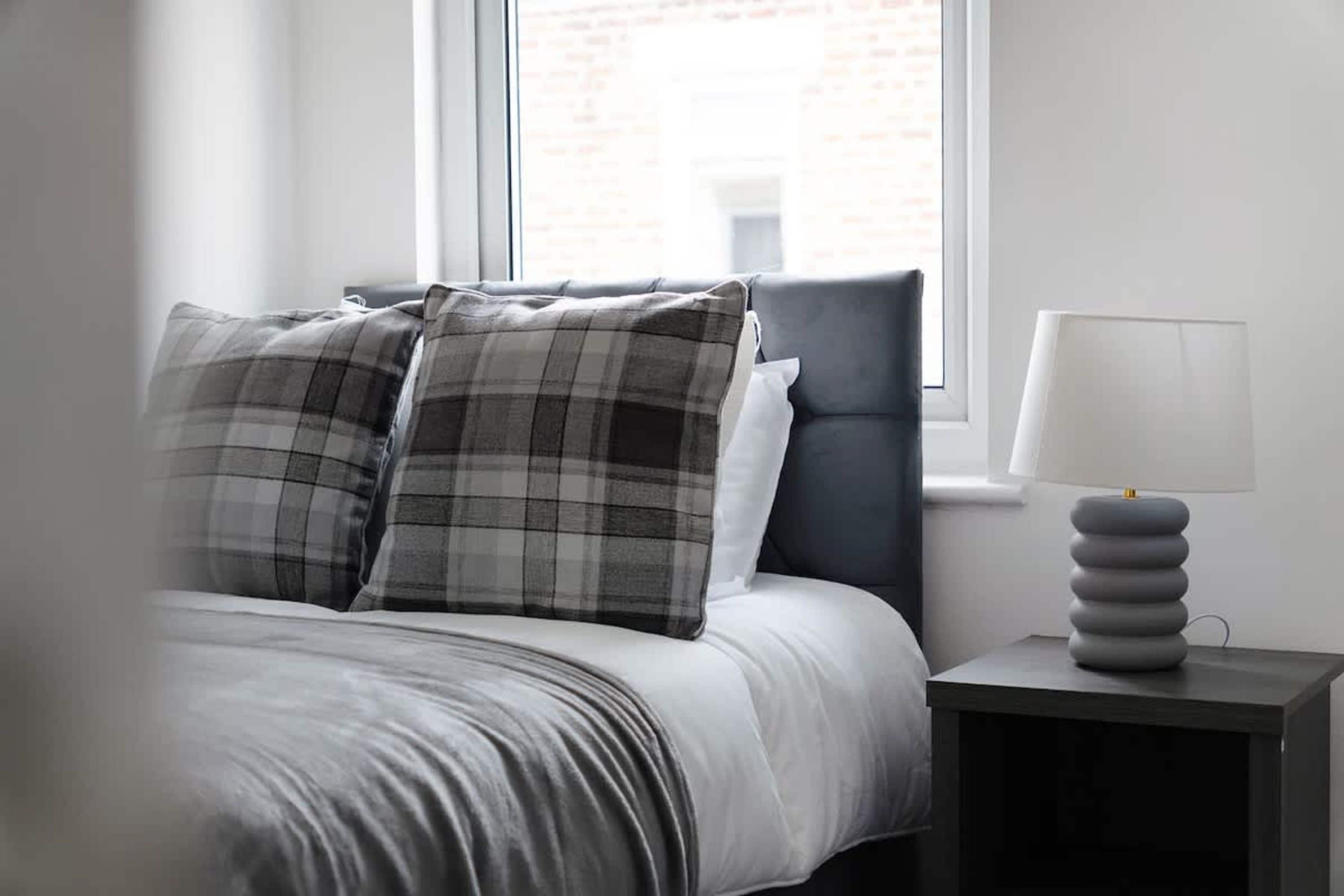A bed with a grey and white checkered pillow, a grey pillow, and a grey headboard. There is a bedside table with a lamp and a stack of books.
