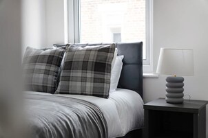 Una cama con un cojín a cuadros gris y blanco, un cojín gris y un cabecero gris. Hay una mesita de noche con una lámpara y una pila de libros.