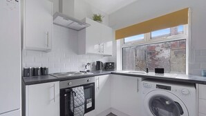 Una cucina compatta con mobili bianchi, forno integrato, lavatrice e finestra con vista su un muro in mattoni.