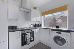 A compact kitchen with white cabinets, a built-in oven, a washing machine, and a window with a view of a brick wall. - Family Coastal Three Bedroom House Parking and Wifi (Saltburn-by-the-Sea)