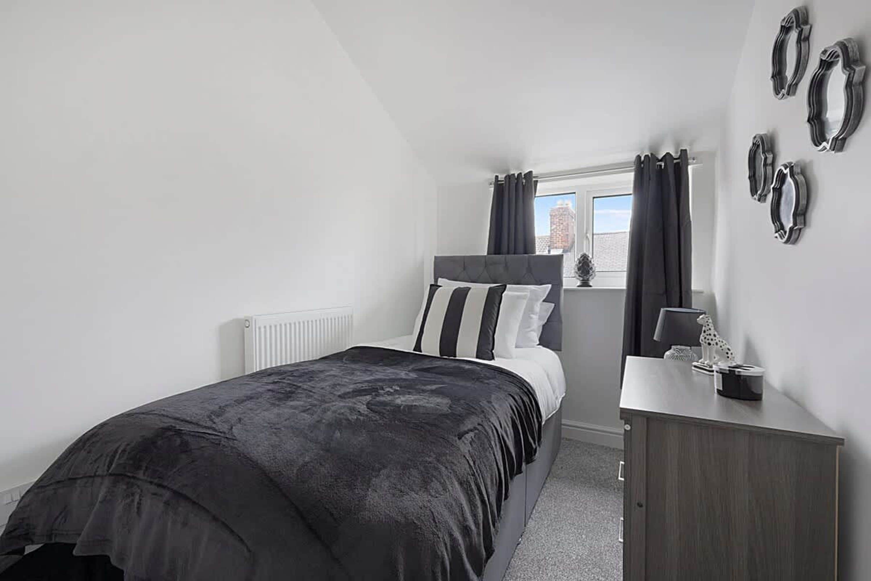 A bedroom with a bed, a bedside table, a dresser, a window with curtains, and a radiator.