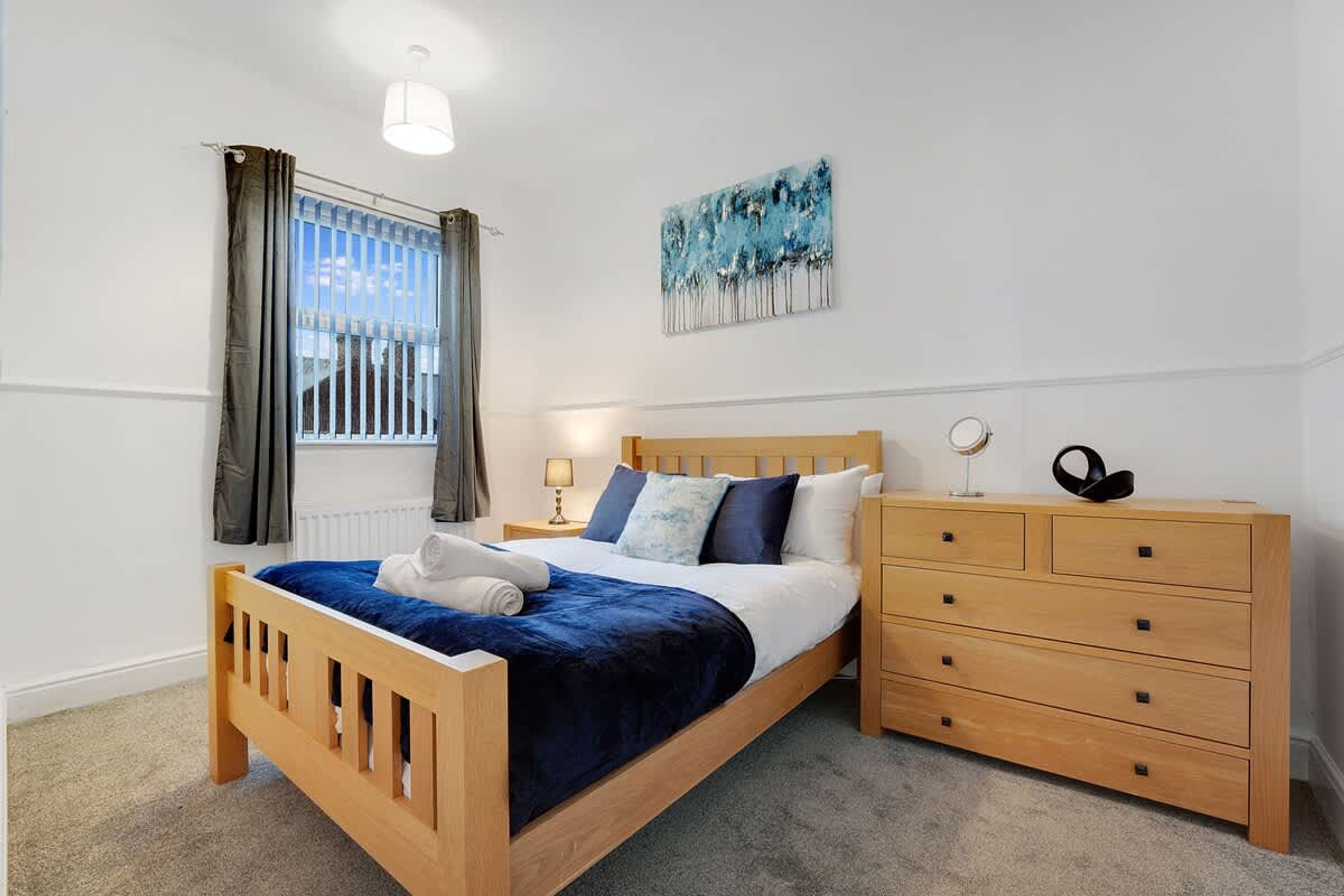 A bedroom with a wooden bed, a dresser, a window with curtains, a painting on the wall, and a lamp on the nightstand.