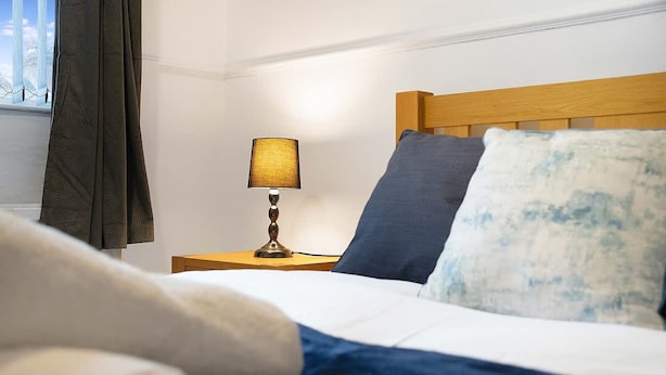 A neatly made bed with a blue blanket and pillows, a bedside lamp, and a window with curtains. - 2BR in Dundee Free Wifi and Indoor Fireplace (Darlington)