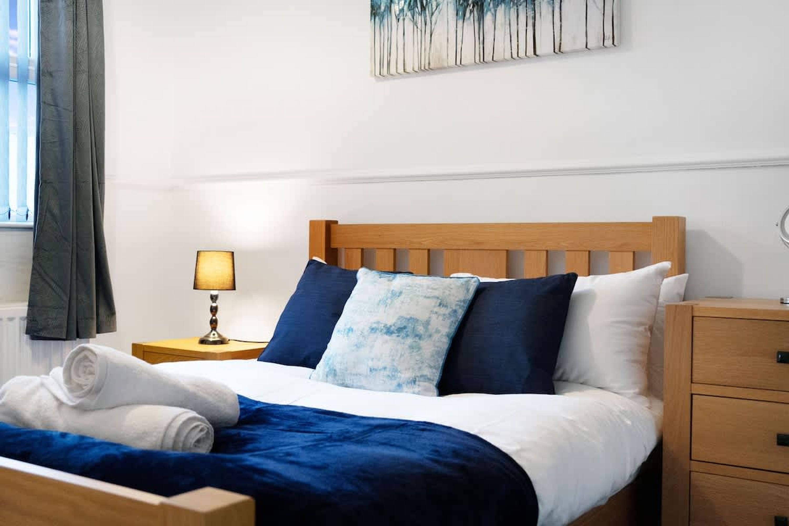 Een opgemaakt bed met een blauw-wit dekbed, een houten hoofdbord en een nachtkastje met een lamp.