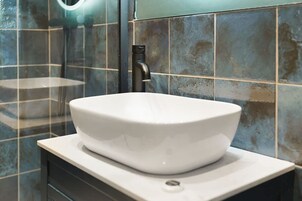 Un baño con un lavabo blanco tipo pedestal, baldosas oscuras y una mampara de vidrio.