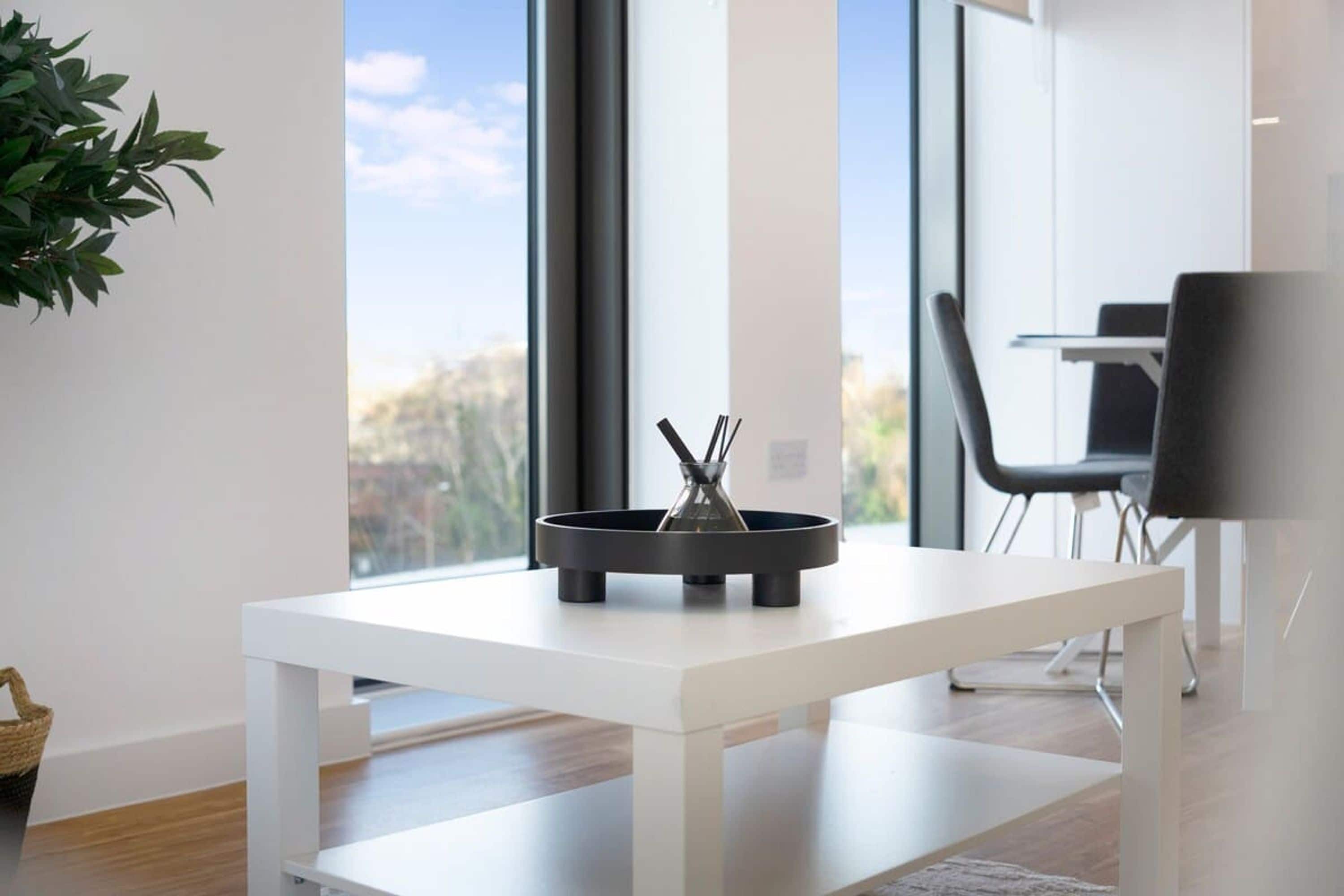 Una habitación moderna y minimalista con una mesa blanca, un objeto negro sobre la mesa y vistas al exterior a través de grandes ventanales.