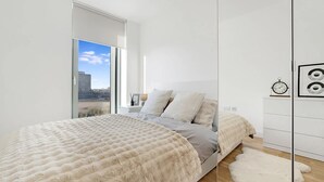 Ein modernes Schlafzimmer mit einem großen Bett, Blick auf die Stadt durch eine Glastür und einer weißen Wand mit einer Uhr.