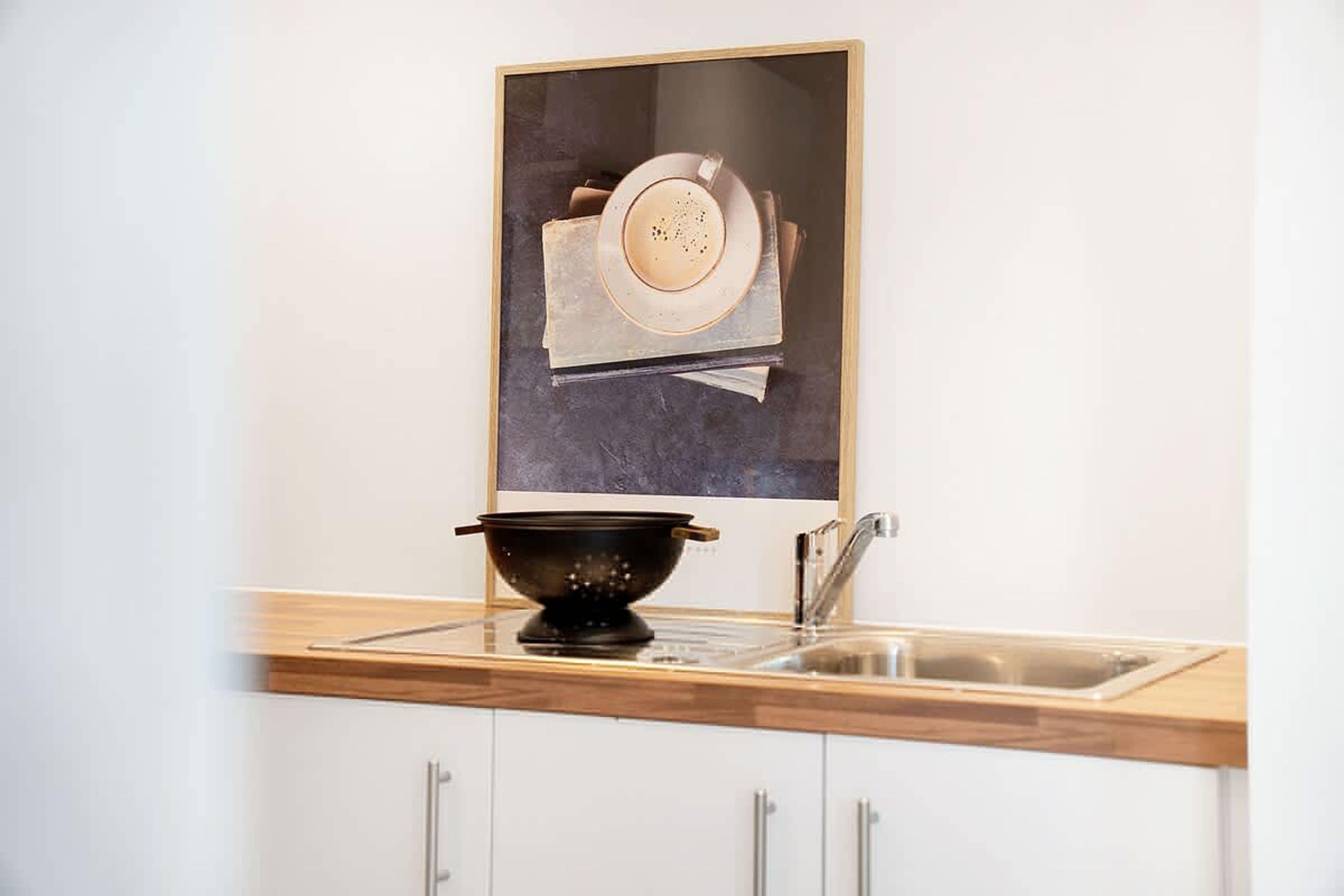 Una cocina con encimera de madera, un colador negro, un lavamanos de acero inoxidable y un cuadro enmarcado sobre el colador.