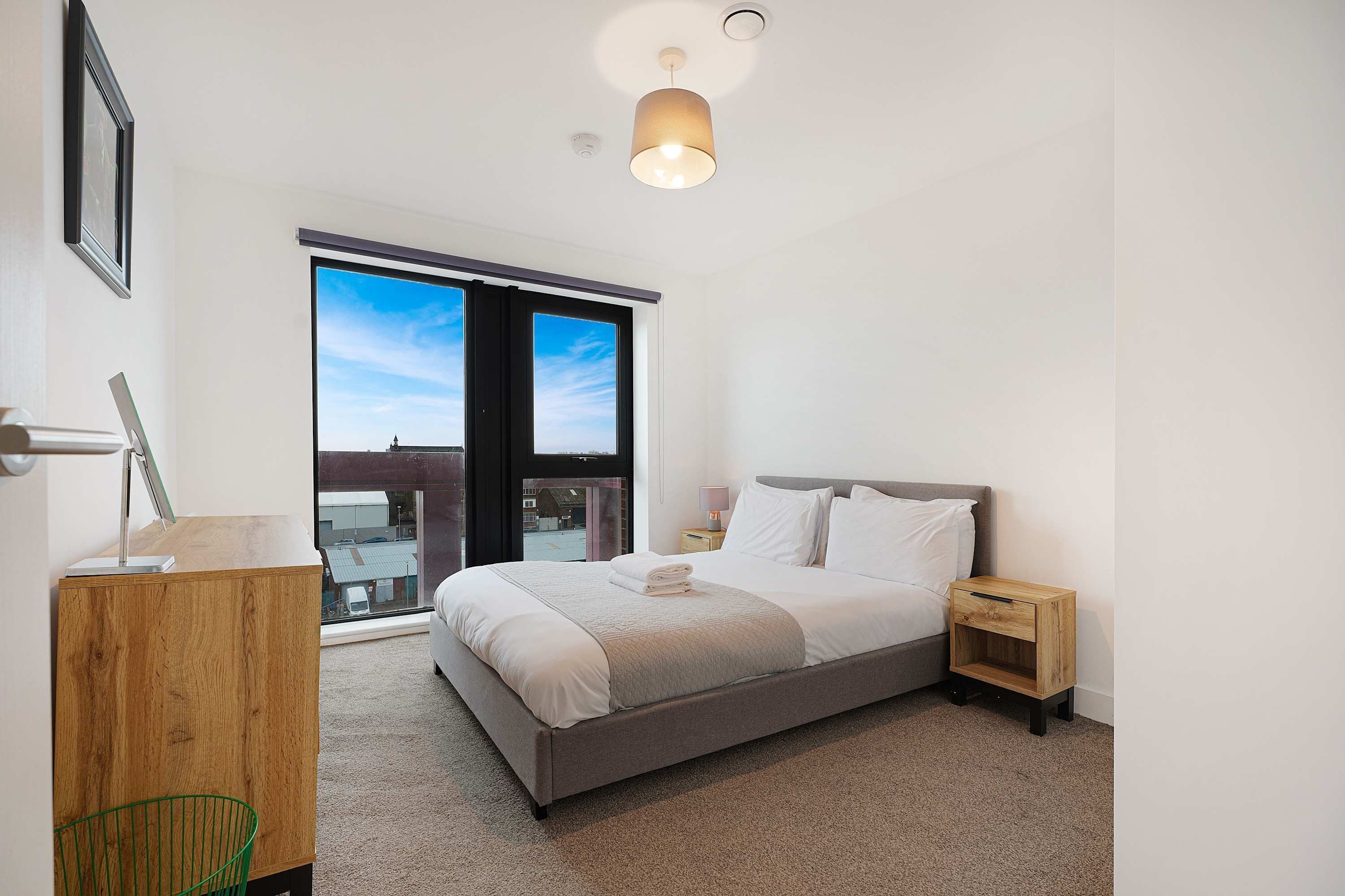 A modern bedroom with a large bed, a bedside table, a lamp, and a view of the sky through a window.