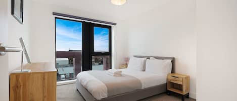 A modern bedroom with a large bed, a bedside table, a lamp, and a view of the sky through a window.