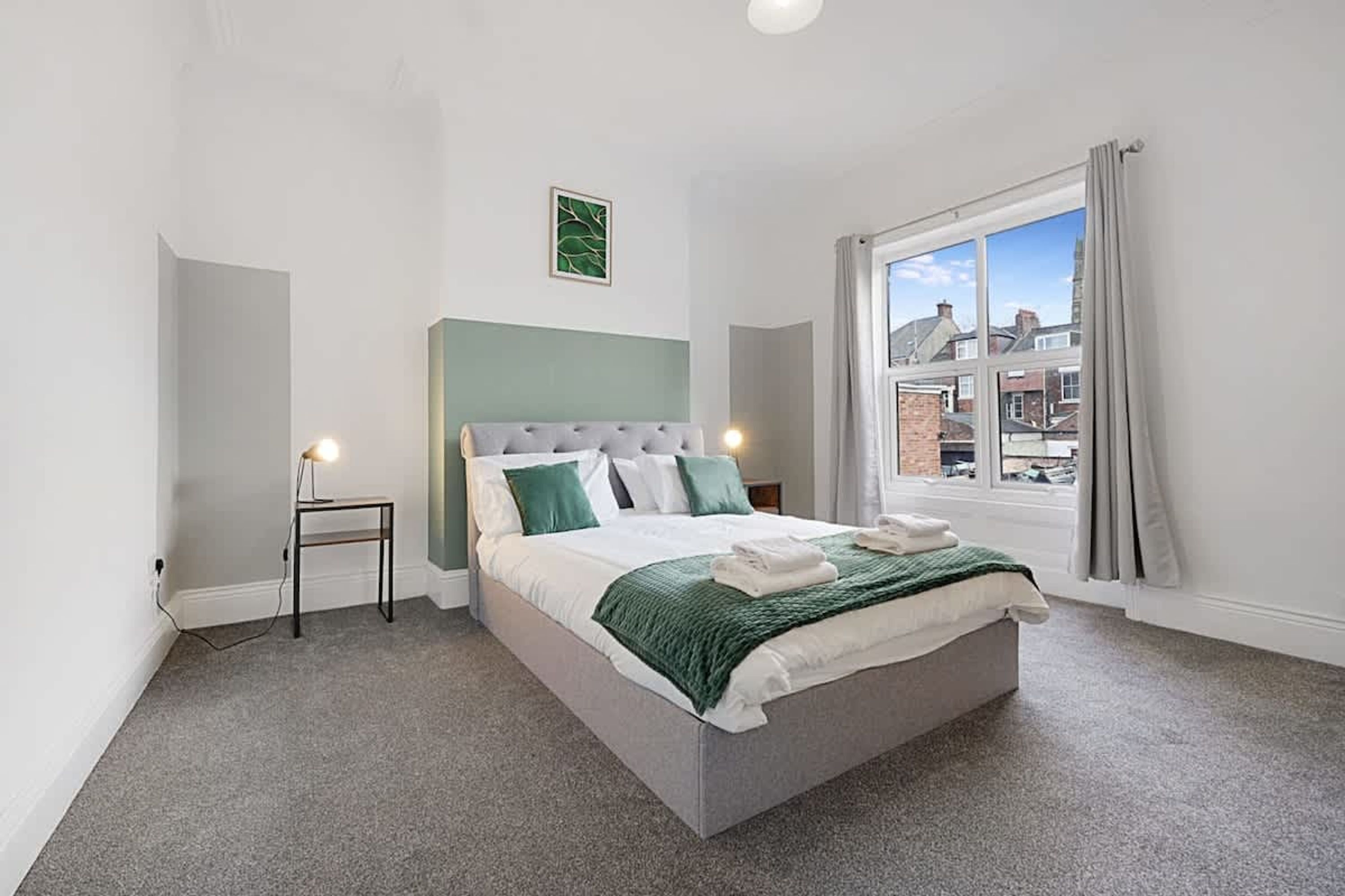 Une chambre à coucher comprenant un lit, une table de chevet, une lampe, une fenêtre donnant sur des immeubles et une œuvre d’art murale de couleur verte.