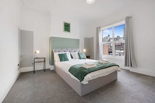 Un dormitorio con cama, mesita de noche, lámpara, ventana con vista a edificios y una obra de arte mural verde.