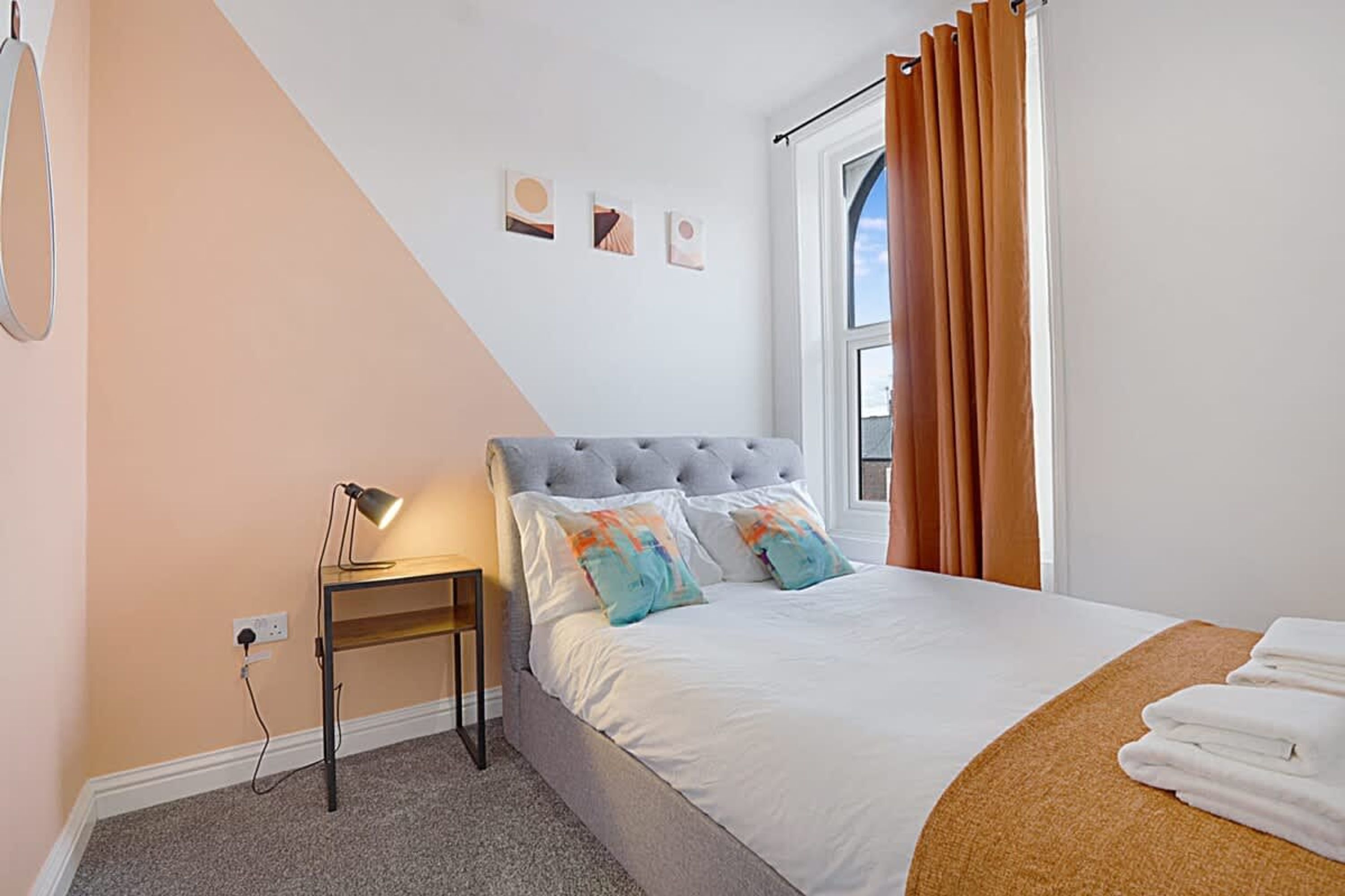 Une chambre à coucher avec un lit, une table de chevet avec une lampe, une fenêtre avec des rideaux et un mur orné de trois tableaux.