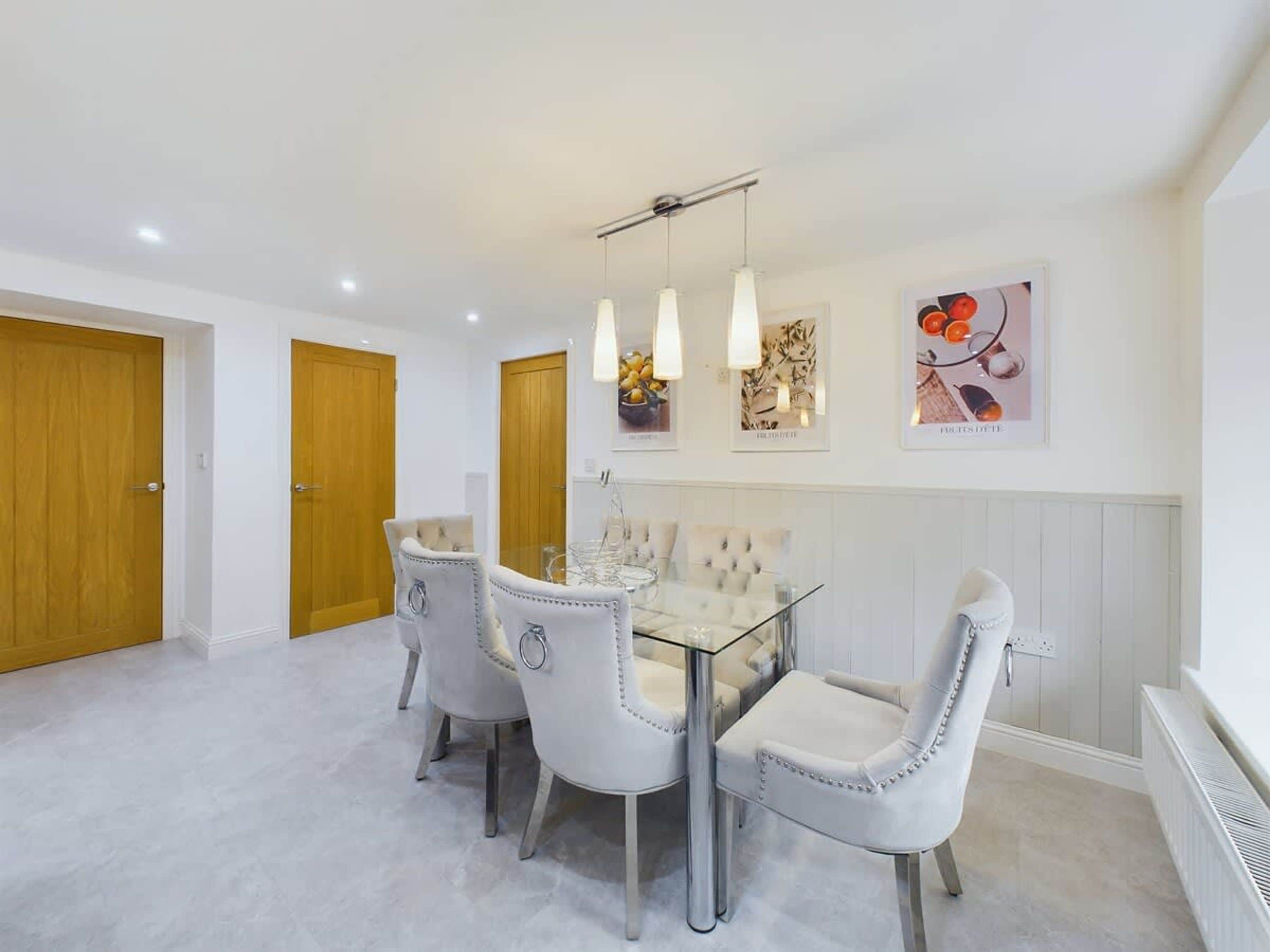 Un comedor con una mesa de vidrio y cuatro sillas tapizadas, puertas de madera y una obra de arte en la pared.