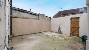 Un espacio exterior estrecho con suelo de hormigón, una puerta de madera y una pared de superficie rugosa.