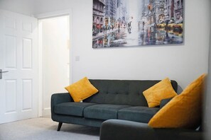 Ein Zimmer mit einem dunkelgrünen Sofa, zwei senffarbenen Kissen, einer weißen Tür und einem Stadtbild an der Wand.