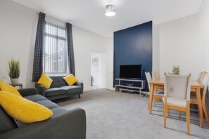 En moderne stue med en grå sofa, gule puder, et fladskærms-tv, et spisebord med stole og et stort vindue med gardiner.