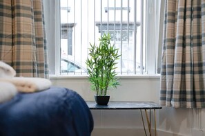 Ein Zimmer mit einem Bett, einem kleinen Tisch mit einer Topfpflanze und einem Fenster mit Vorhängen.