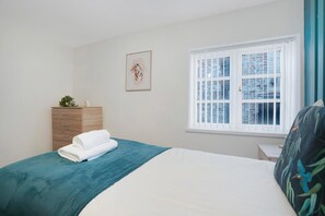 Ein Schlafzimmer mit einem Bett, einem Schminktisch, einem Fenster und einem gerahmten Bild an der Wand.