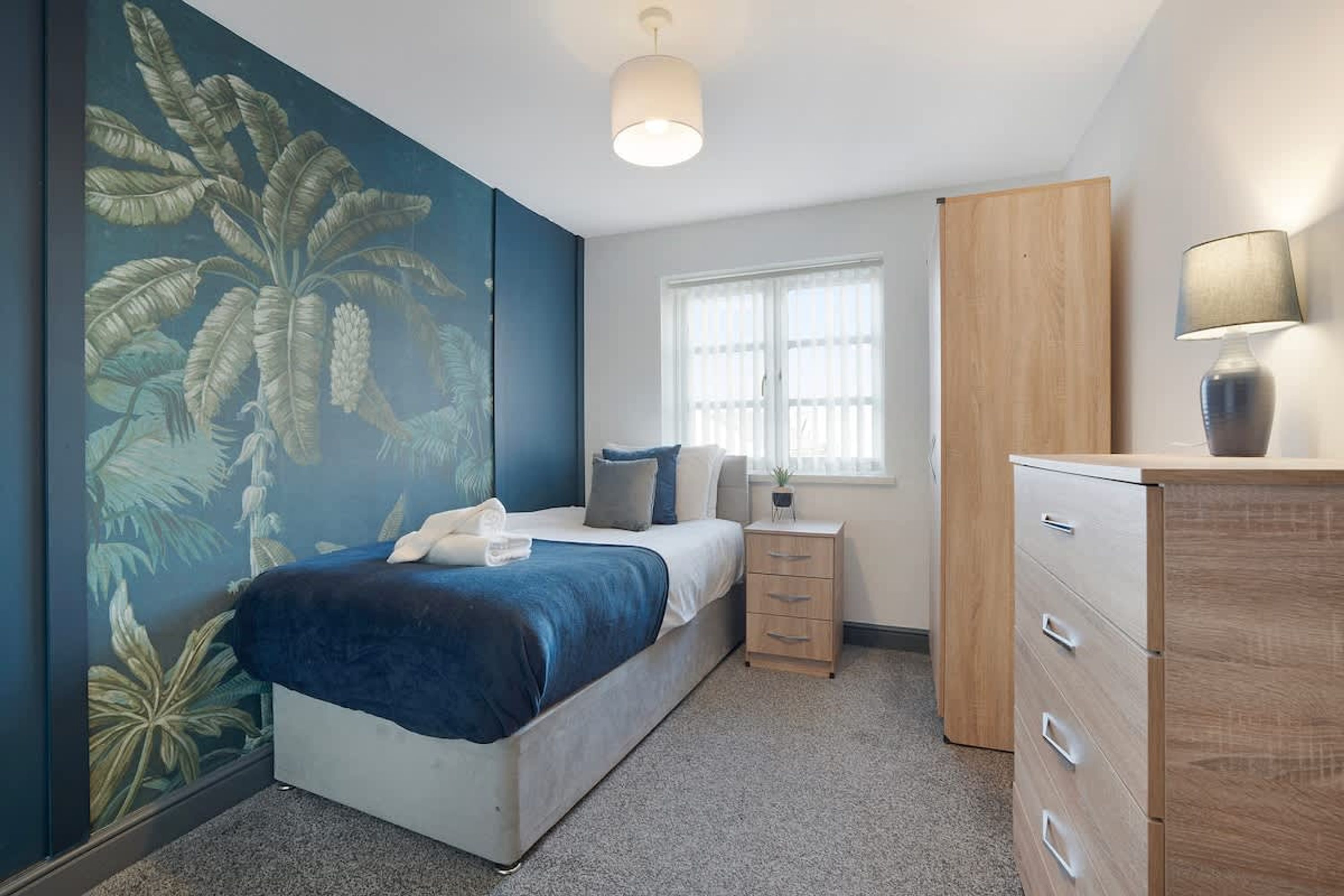 A bedroom with a bed, a nightstand, a dresser, a window, and a wall with a leaf pattern.