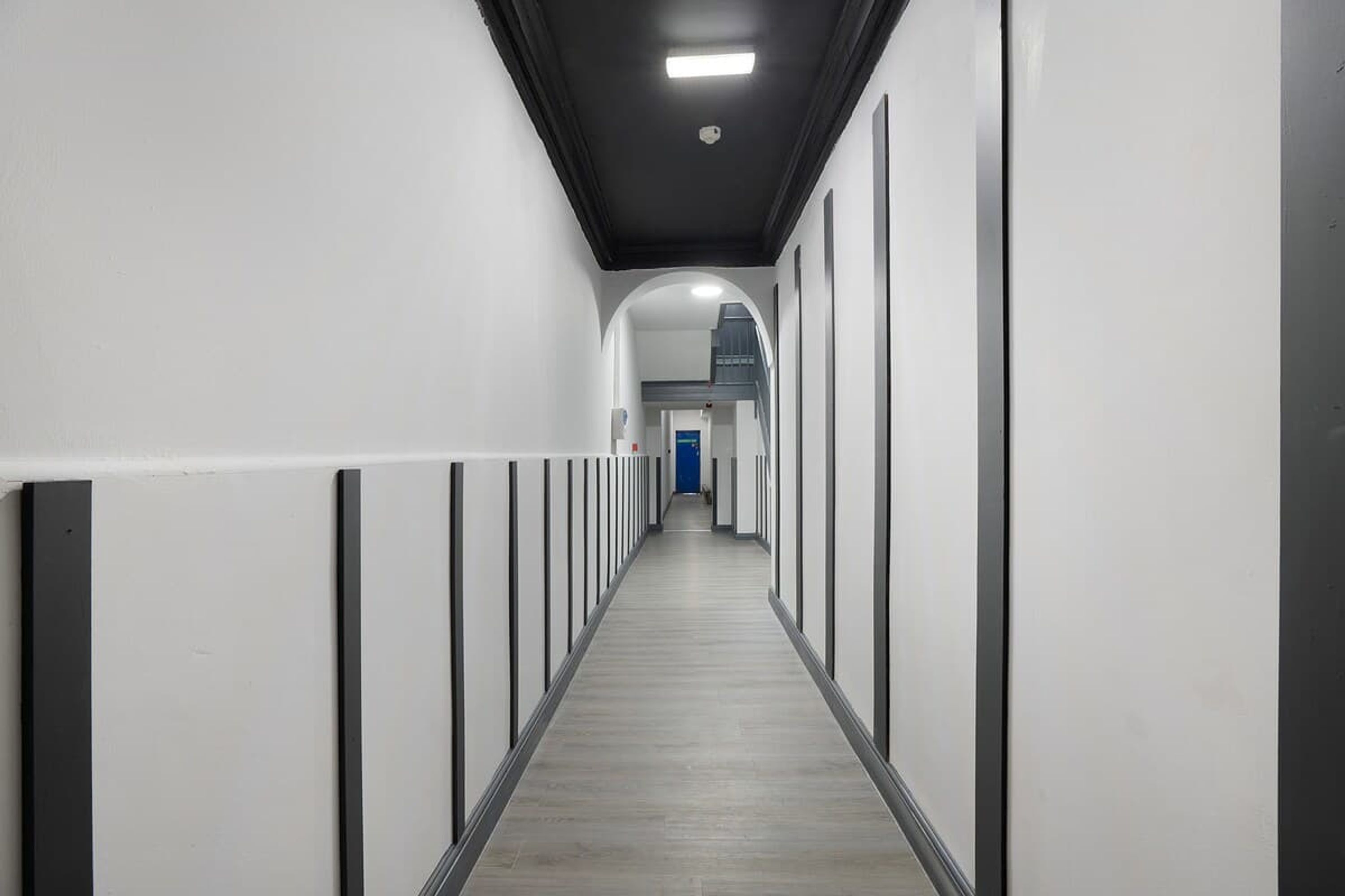 A long, narrow hallway with white walls and a grey floor, leading to a blue door at the end.