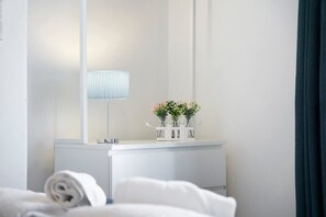 Ein Hotelzimmer mit weißem Schminktisch, einem Spiegel, einer Lampe und einer Vase mit Blumen.