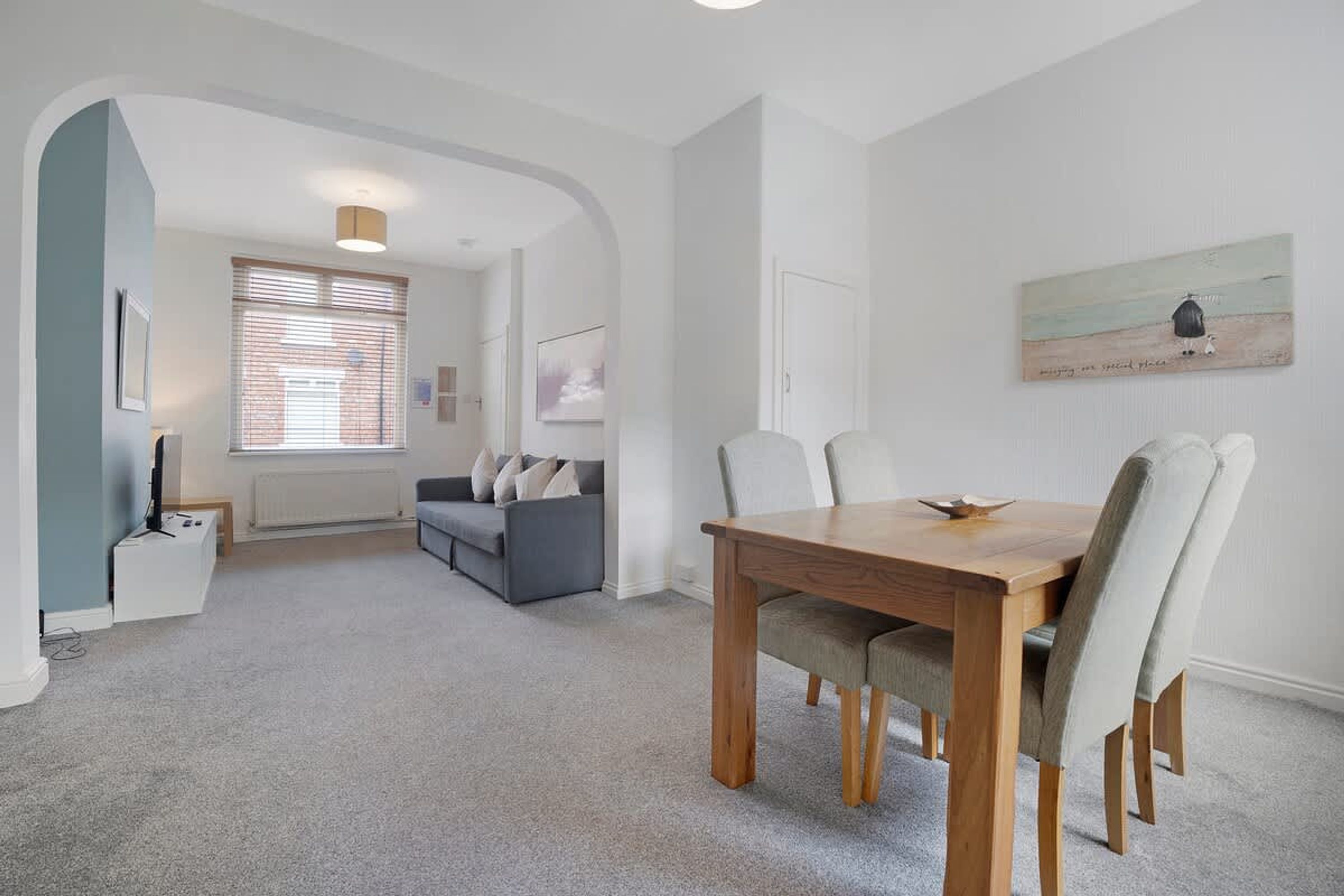 A modern living room with a grey sofa, a dining table with chairs, and a painting on the wall.