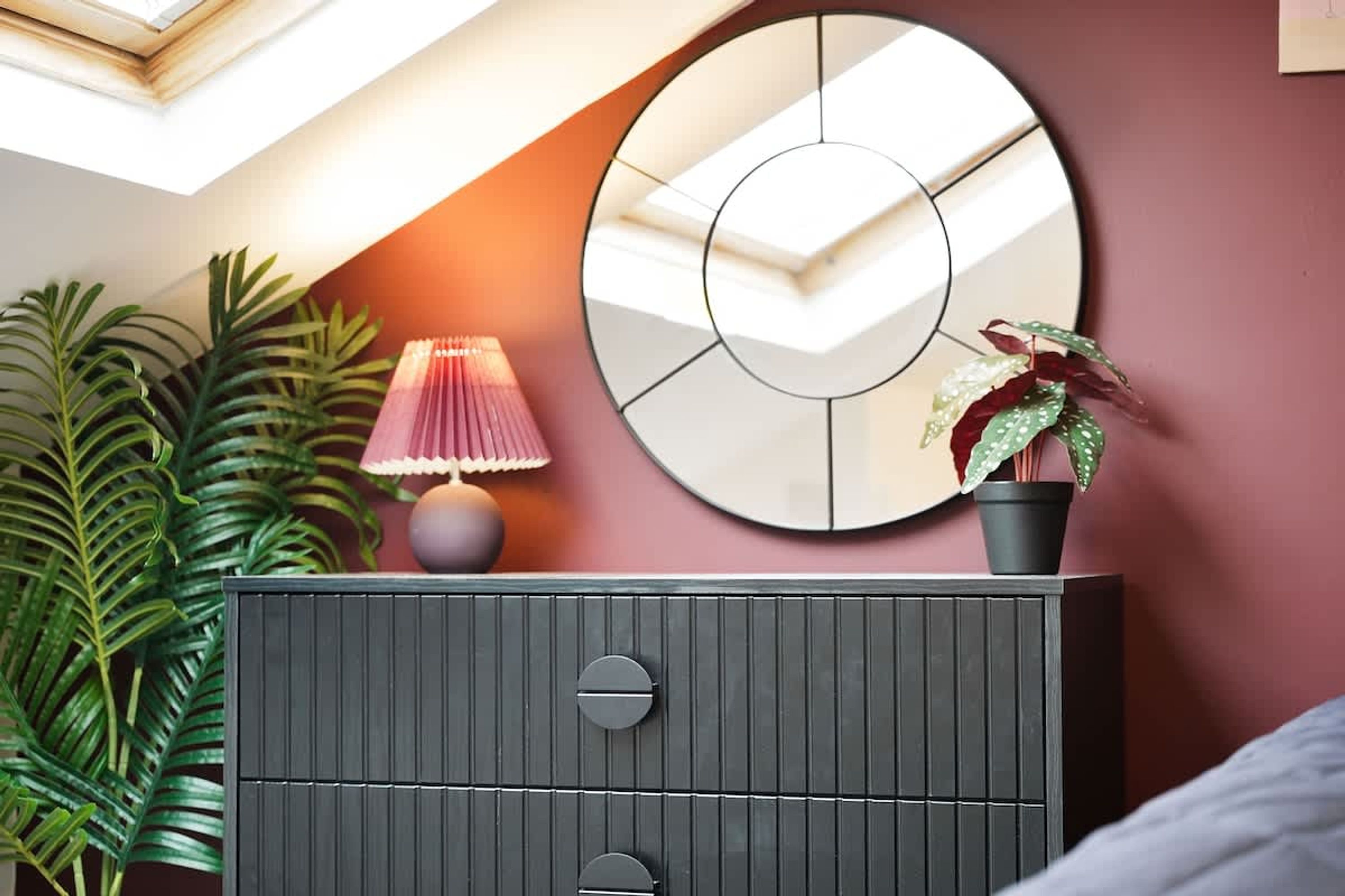 A room with a dark grey dresser, a round mirror, a pink lamp, and a potted plant.