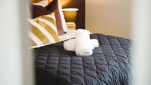 Ein ordentlich bezogenes Bett mit einer dunklen Decke, einem aufgerollten weißen Handtuch und einem braunen Kopfkissen.