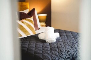 A neatly made bed with a dark quilt, a rolled white towel, and a brown pillow. - Modern 4BR With Easy Access to Town and Station (Darlington)