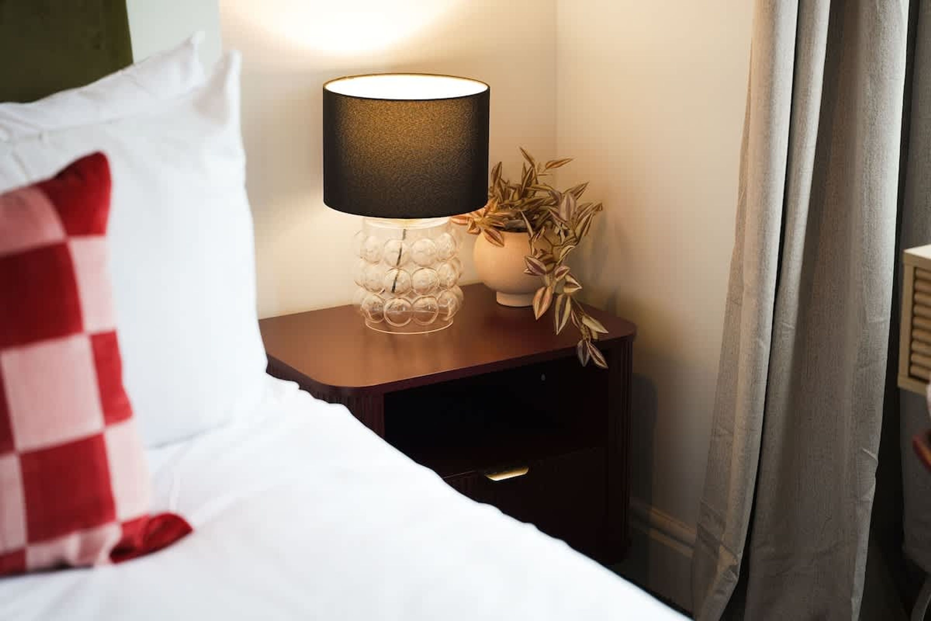 Un lit bien fait, avec une literie blanche, un coussin à carreaux rouges et blancs, une lampe de chevet avec un abat-jour texturé et une petite plante en pot posée sur une table de chevet en bois.