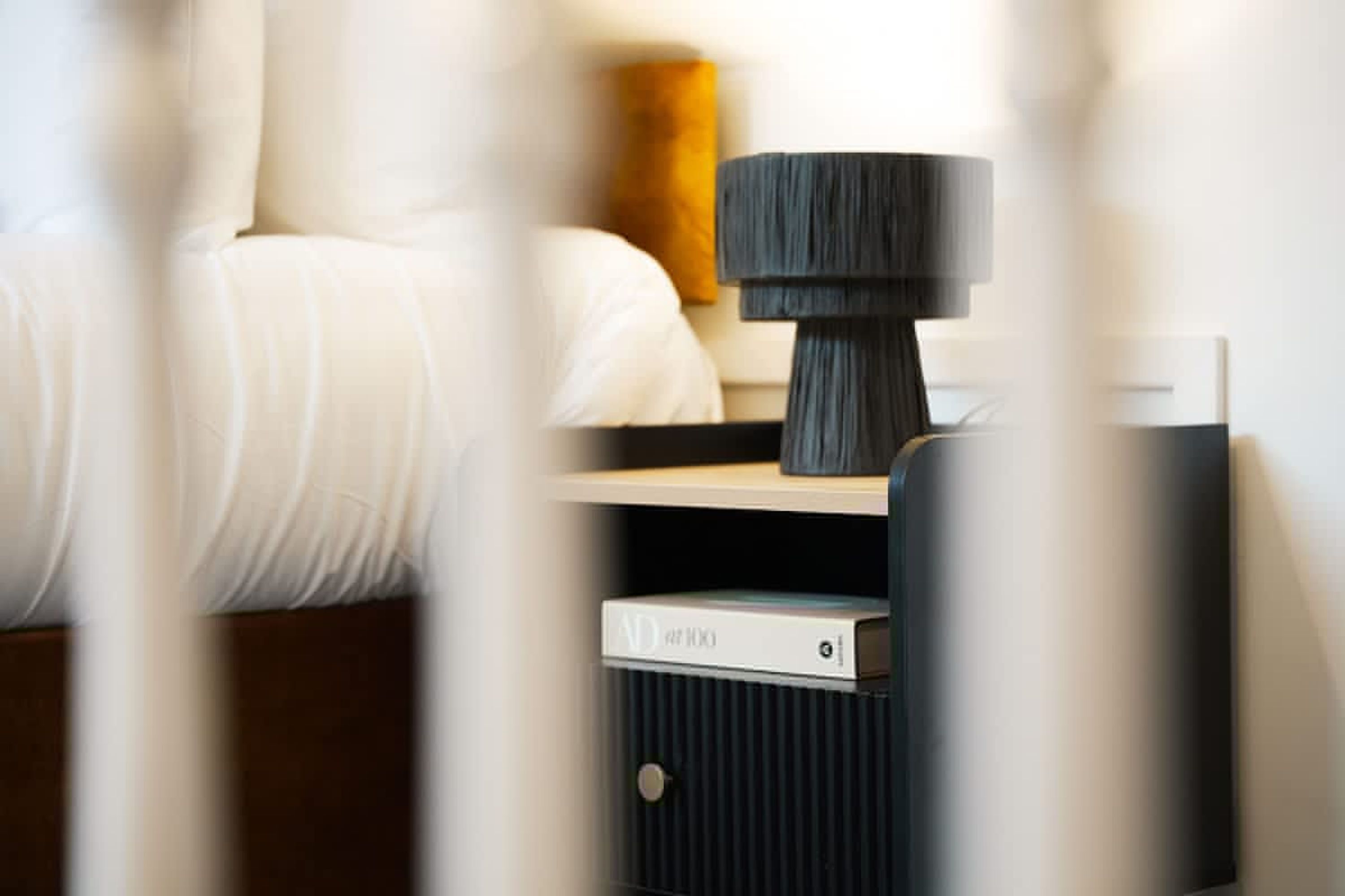 A neatly made bed with white linens, a bedside table with a lamp, and a small electronic device on the nightstand.