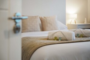 Ein ordentlich bezogenes Bett mit einer beigen Decke und zwei Handtüchern, ein Nachttisch mit einer Lampe und eine Tür mit Griff.