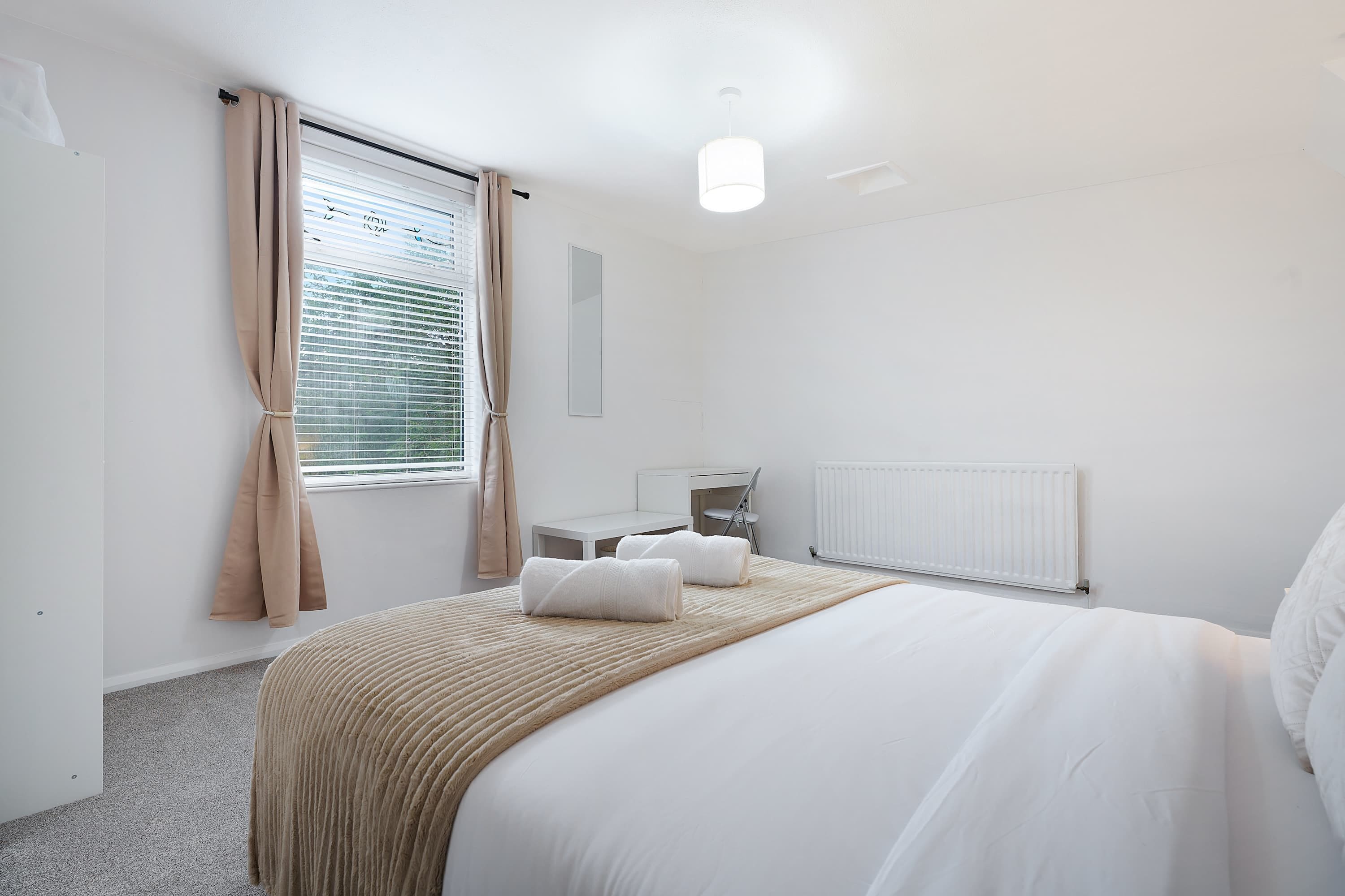 A hotel room with a bed, a desk, a chair, a radiator, and a window with curtains.