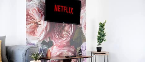 Una sala de estar con un televisor de pantalla plana montado sobre un papel tapiz floral, un sofá gris, una pequeña mesa de centro de madera, una planta en maceta y una mesita de noche con un control remoto.