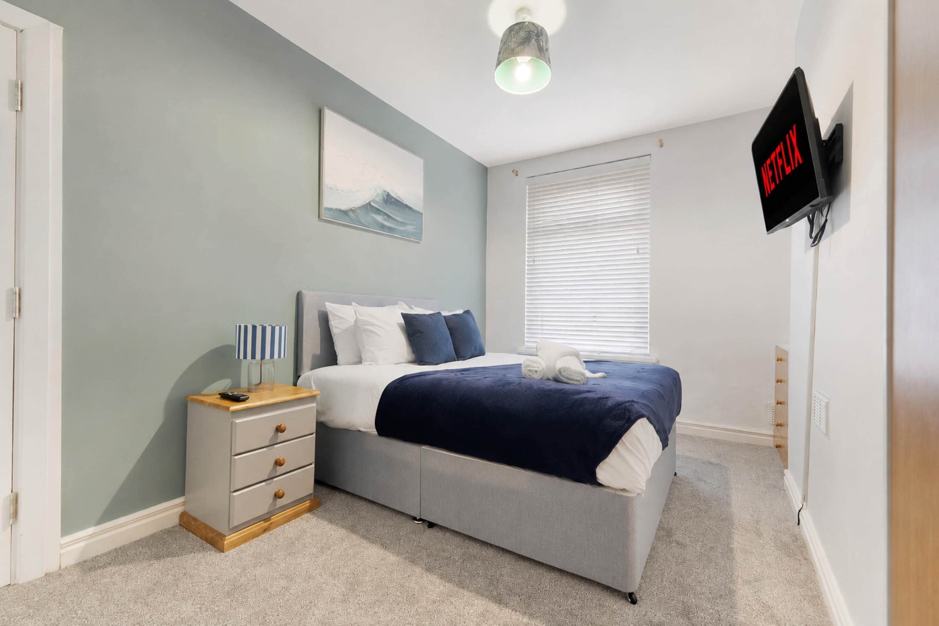 A bedroom with a bed, bedside table, lamp, artwork, window, and a TV mounted on the wall.